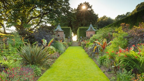 A long stretch of grass in a garden with borders of red flowers leading up to steps with two brick gazebos either side, a low sun shining through the trees in the background.